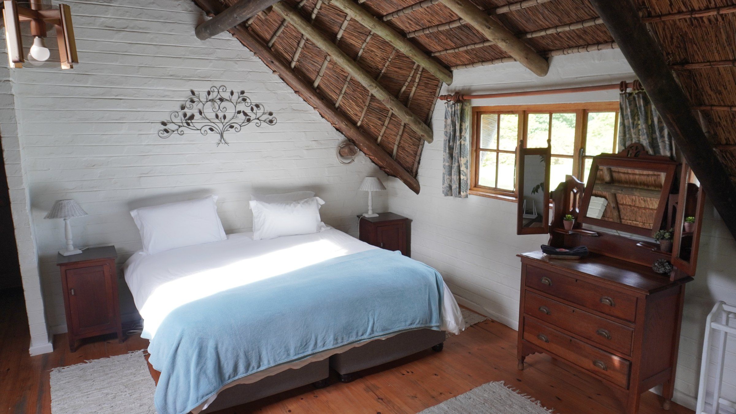 The loft bedroom under the thatch, the bed made up with a blue throw beside an old wooden dresser